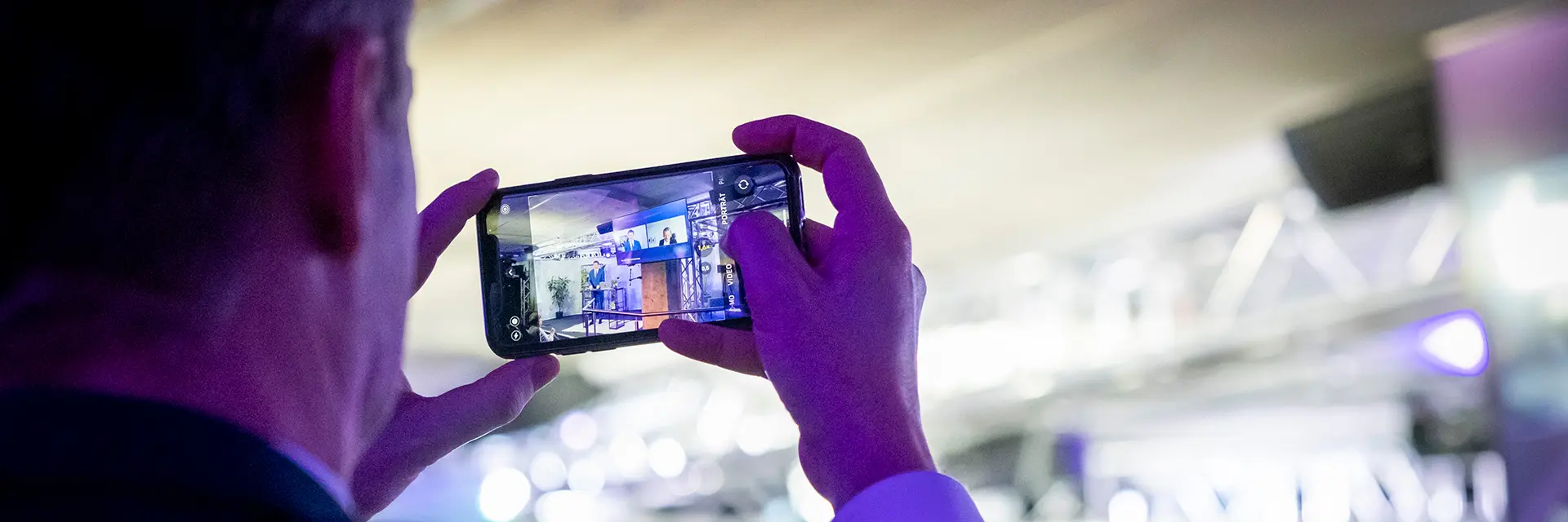 Person fotografiert mit dem Smartphone eine Präsentation auf einer Bühne in einem beleuchteten Veranstaltungsraum.