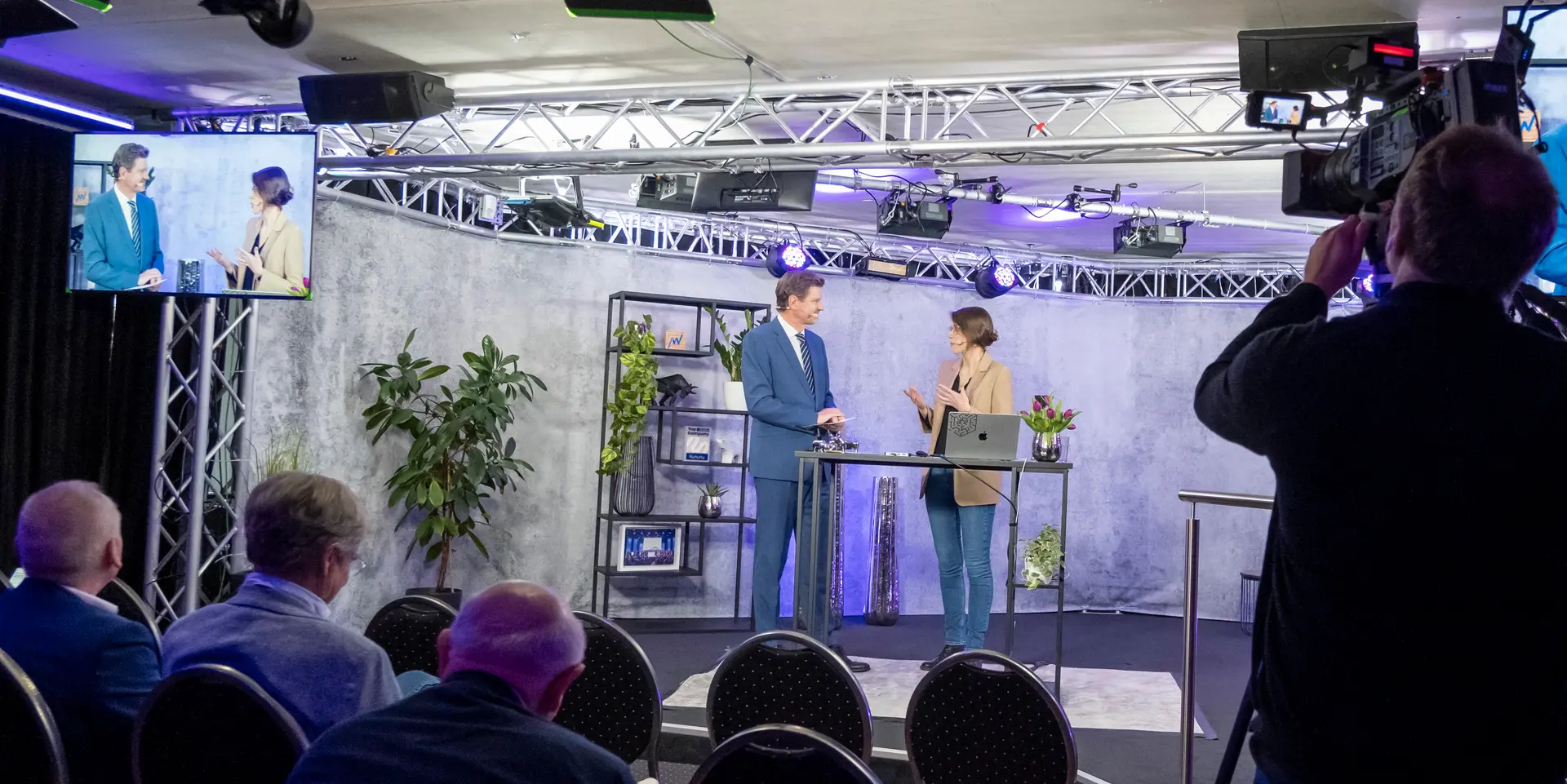 Zwei Personen präsentieren auf einer kleinen Bühne in einem Studio mit Kameras, Scheinwerfern und sitzendem Publikum.