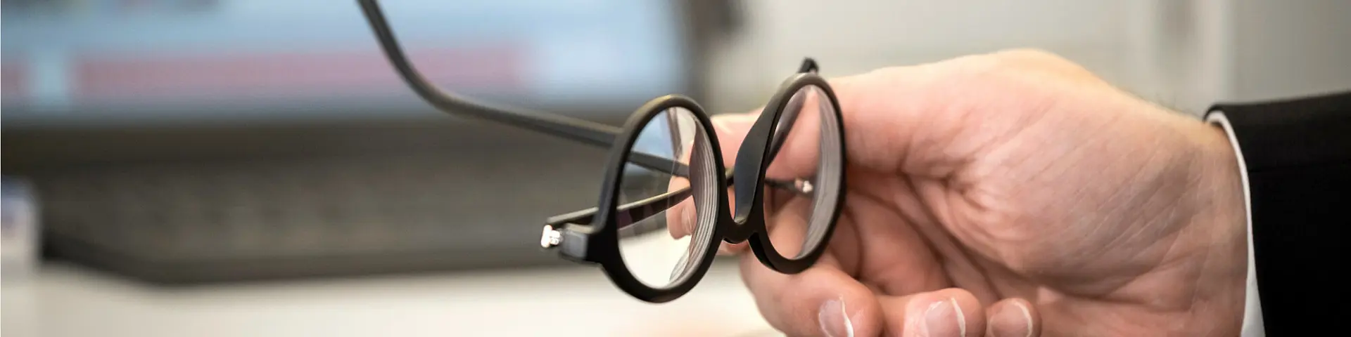 Eine Hand hält eine runde, schwarze Brille vor einem unscharfen Bildschirm im Hintergrund.