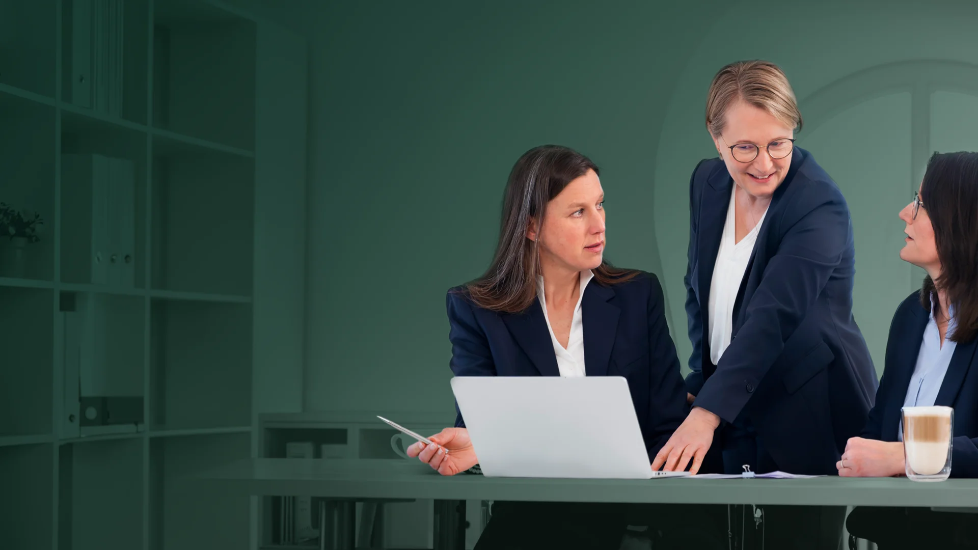 Drei Frauen sitzen in einem weißen Büro an einem Schreibtisch. Die Frau in der Mitte zeigt auf einen Laptop.