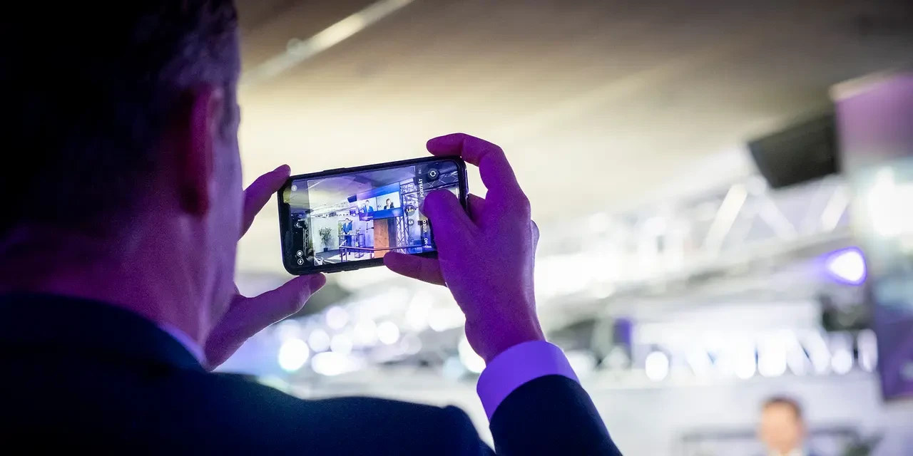 Person fotografiert mit dem Smartphone eine Präsentation auf einer Bühne in einem beleuchteten Veranstaltungsraum.