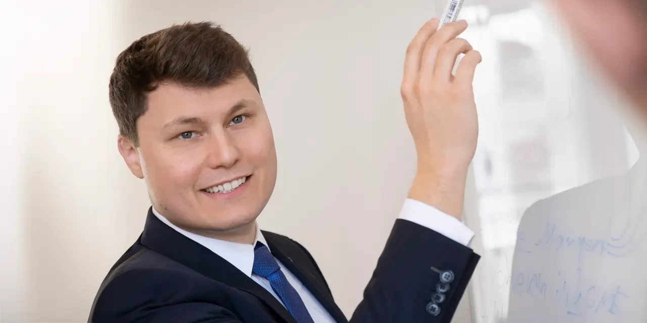 Ein Mann im Anzug schreibt lächelnd auf ein Whiteboard.
