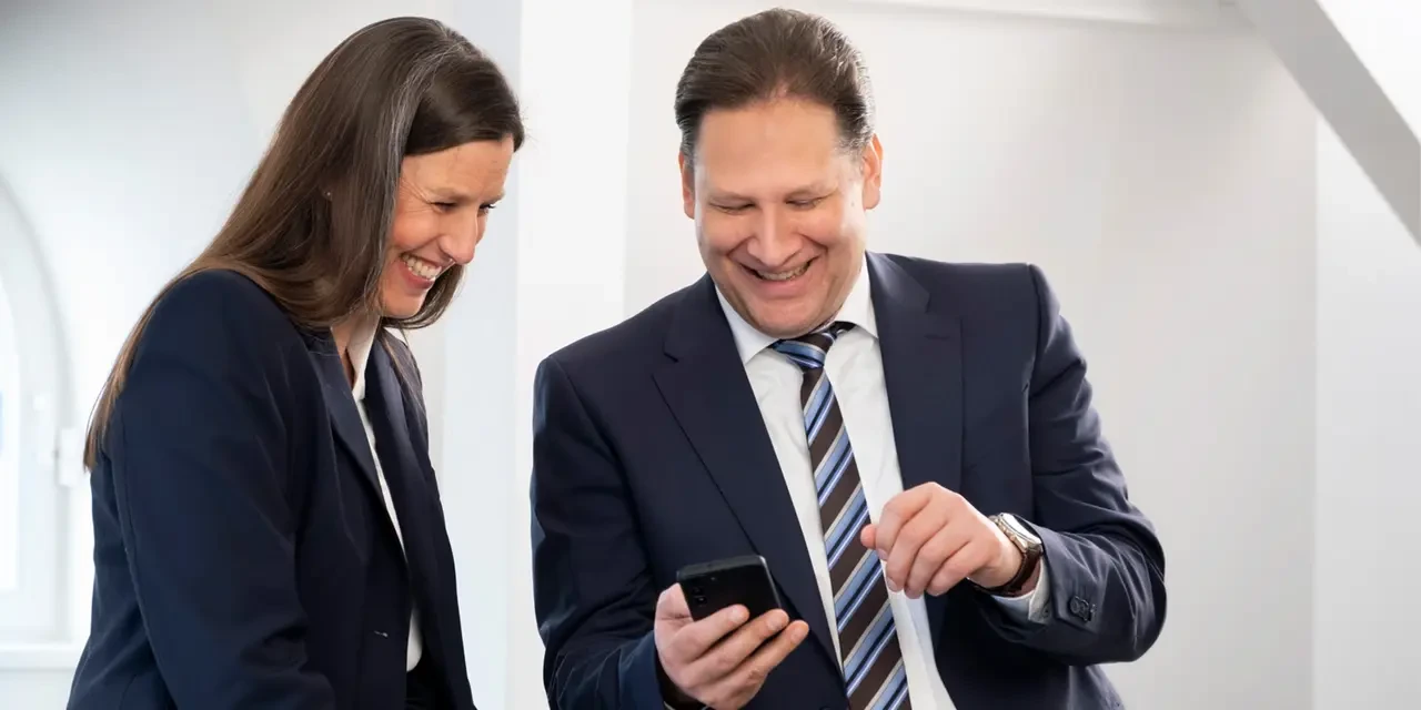 Ein lächelnder Mann zeigt einer Frau auf seinem Smartphone etwas, während sie gemeinsam darauf schauen.