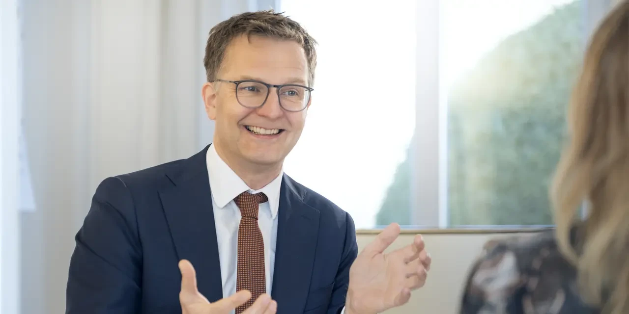 Lächelnder Berater mit Brille erklärt einer Gesprächspartnerin etwas mit lebhafter Gestik in einem hellen Büro 