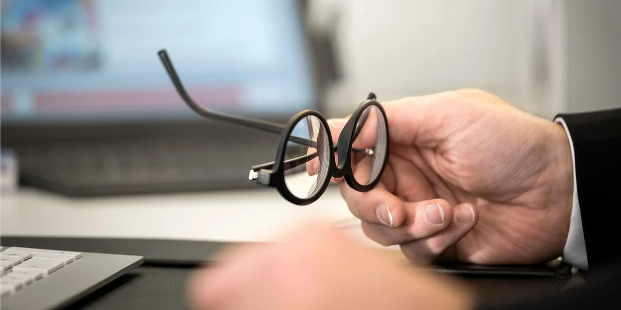 Eine Hand hält eine runde, schwarze Brille vor einem unscharfen Bildschirm im Hintergrund