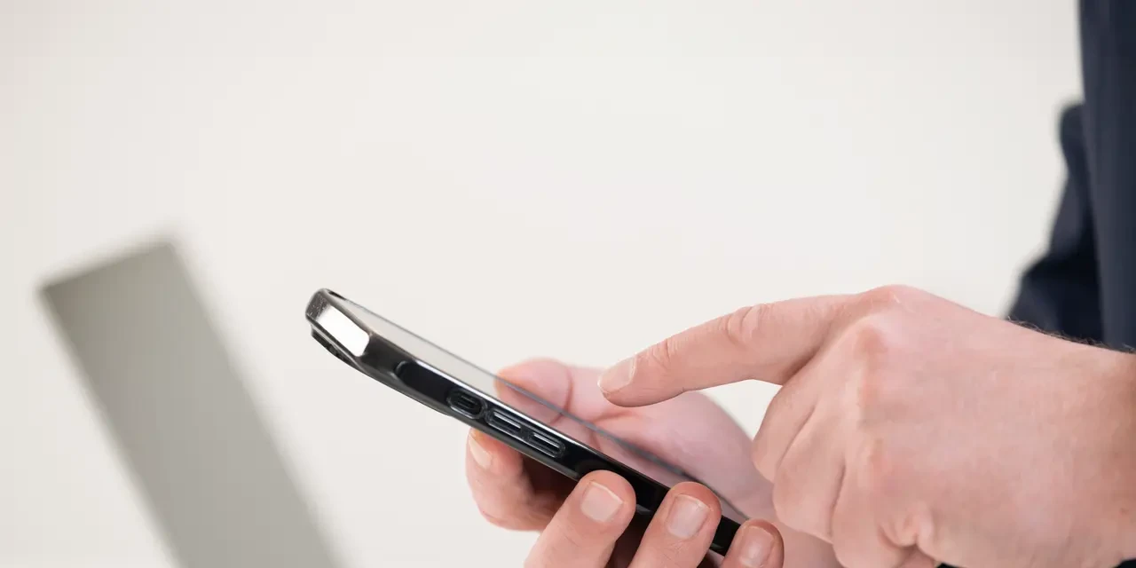 Hand bedient ein Smartphone, im Hintergrund unscharf ein aufgeklappter Laptop 
