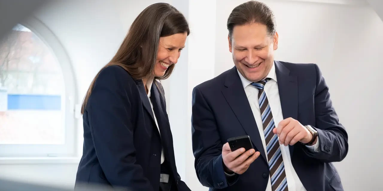 Ein lächelnder Mann zeigt einer Frau auf seinem Smartphone etwas, während sie nebeneinander stehen.