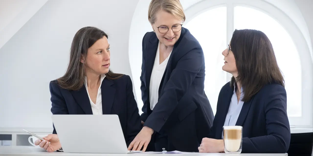 Drei Frauen sitzen in einem weißen Büro an einem Schreibtisch. Die Frau in der Mitte zeigt auf einen Laptop.