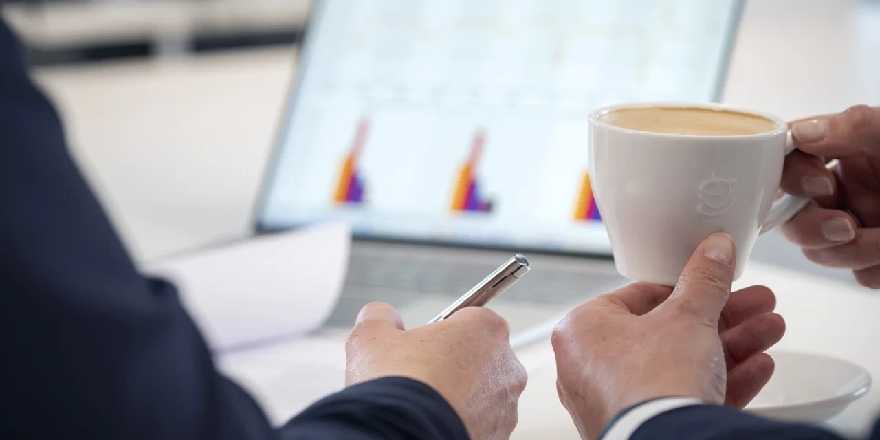Zwei Personen im Büro: Eine hält eine Tasse Kaffee, die andere einen Stift; im Hintergrund ein Laptop mit Diagrammen.