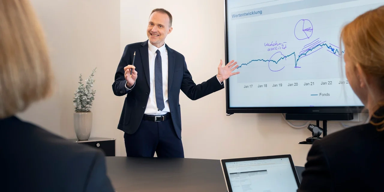 Ein Mann hält eine Präsentation vor einem Bildschirm mit Diagramm, während zwei Personen zuhören.