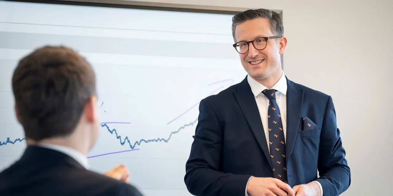 Zwei Männer im Anzug im Gespräch vor einem Whiteboard mit eingezeichnetem Diagramm; der Mann im Vordergrund ist unscharf, der andere lächelt in die Kamera.